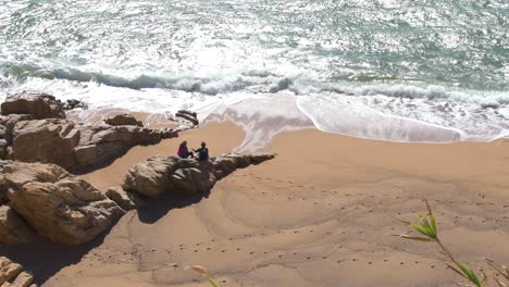 Vista-Aérea-De-Una-Pareja-Sentada-En-Las-Rocas-Mirando-Las-Olas-Del-Mar-En-La-Playa.