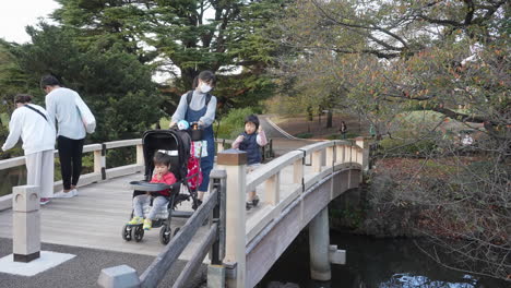 Mutter-Schiebt-Baby-Auf-Kinderwagen,-Während-Sohn-Neben-Ihr-Auf-Einer-Hölzernen-Fußgängerbrücke-Im-Nationalpark-Und-Garten-Shinjuku-Gyoen-In-Tokio,-Japan,-Läuft