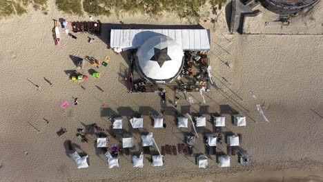 AERIAL:-Top-View-Shot-of-White-Tents-on-a-Sandy-Nida-Beach-with-Waving-Flags-in-Wind-and-Long-Shadows