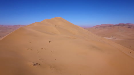 Aerial-landscape-of-Chile's-Atacama-desert,-rising-over-a-large-sand-dune-to-reveal-the-horizon,-bright-blue-sky