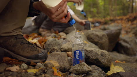 A-Hiker-Filtering-Clean-Water-into-a-Water-Bottle-Next-to-a-Stream-in-a-Forest-with-Beautiful-Fall-Colors,-Slow-Motion