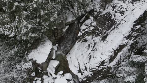 Agua-Que-Fluye-Rápidamente-Sobre-Una-Cascada-Mientras-Los-Copos-De-Nieve-Blanca-Giran-Entre-Los-Pinos-Y-Las-Rocas-En-Una-Escena-De-Invierno