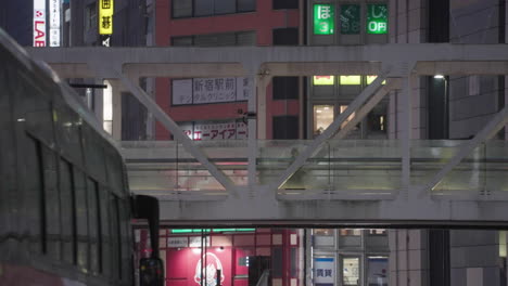 Mujer-Adulta-Caminando-Sobre-Un-Puente-Peatonal-En-Una-Noche-Lluviosa-Desde-La-Acera-Con-El-Autobús-Urbano-En-Shinjuku,-Tokio,-Japón