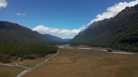 Cars-travelling-through-a-beautiful-mountain-valley-with-river-and-large-open-grass-fields-in-Fiordland-Southland,-New-Zealand