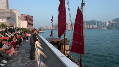 People-gather-at-Victoria-Harbour-waterfront-as-a-wooden-red-sailed-junk-boat-based-on-ancient-Chinese-sailing-ships,-now-used-as-a-touristic-attraction,-is-seen-in-Hong-Kong
