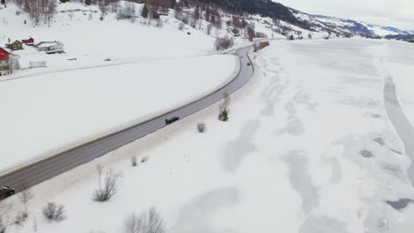 Coches-Conduciendo-Por-Una-Carretera-Sinuosa-Cerca-De-Un-Pequeño-Pueblo-Y-Campos-Nevados-En-Invierno-En-Haugastol,-Noruega