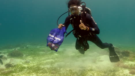 Volunteer-Scuba-Diver-Making-Okay-And-Thumbs-Up-Hand-Gestures-While-Holding-A-Bag-Of-Collected-Garbage-Under-The-Sea