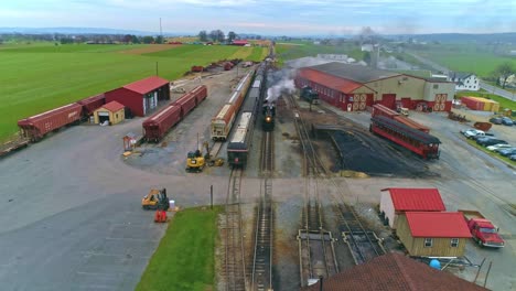 An-Aerial-View-of-an-Antique-Restored-Steam-Train-and-Passenger-Coaches-Approaching-With-Smoke-and-Steam-Pulling-into-the-Yard-and-Station-as-a-Second-Steam-Train-is-Leaving-as-Seen-by-a-Drone