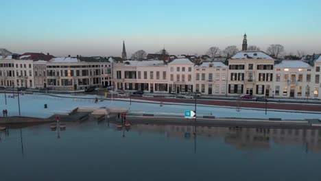 Boulevard-with-snow-view-rotating-aerial-showing-the-full-cityscape-of-Dutch-Hanseatic-medieval-tower-town-Zutphen,-The-Netherlands,-seen-from-the-river-IJssel-in-the-foreground-at-sunset-blue-hour