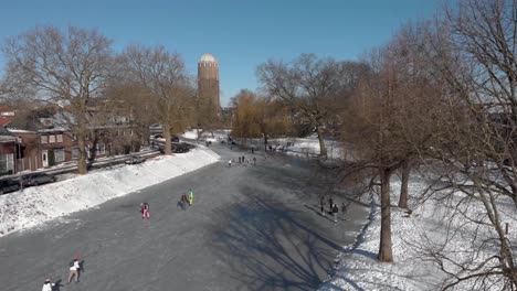 Picturesque-aerial-view-of-people-ice-skating-along-the-curved-frozen-canal-going-through-the-Dutch-city-of-Zutphen-with-shadows-of-barren-trees-and-former-water-tower-in-the-background