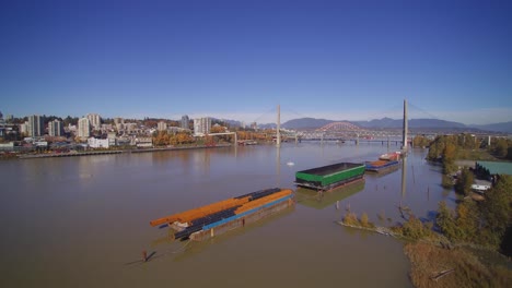 Aerial-4K-Island-in-Fraser-River-with-green-trees-city-of-new-westminster-in-background-blue-sky-bright-sunny-day-boats-moored-and-Patullo-Bridge