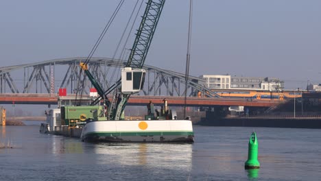 Schiff-Mit-Großem-Kran,-Der-Den-Frachtschiffkorridor-Des-Flusses-Ijssel-Ausbaggert,-Der-Unter-Der-Zugbrücke-Dahinter-Führt,-Mit-Einem-Gelb-blauen-Zug-Der-Niederländischen-Staatsbahn,-Der-Im-Hintergrund-Vorbeifährt