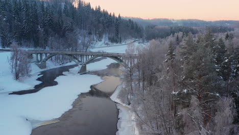 Brücke-über-Den-Gefrorenen-Fluss,-Sonnenuntergang-über-Verschneitem-Winterwald,-4K-Luftaufnahme