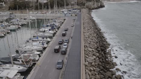 Cars-Driving-On-Promenade-In-Barcelona,-Spain-With-Sailboats-Moored-At-Port