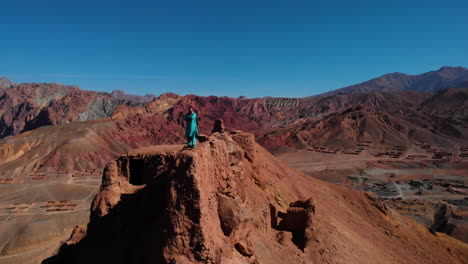 Man-On-Top-Of-Rugged-Cliff-Overlooking-Red-City-In-Bamyan,-Afghanistan---aerial-pullback