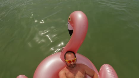 Man-with-yellow-sunglasses-taking-selfie-holding-long-stick-and-camera-while-relaxing-on-floating-inflatable-pink-flamingo