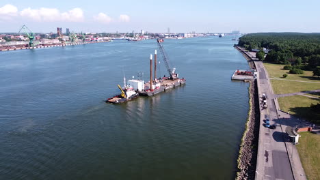 Tugboat-pushing-barge-with-crane-on-top-to-docks-of-Klaipeda-harbor,-aerial-view