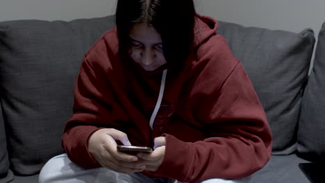 A-teenage-youth-dressed-in-a-hoody-sitting-on-a-sofa-at-home,-the-young-girl-focused-on-her-mobile-phone-as-she-messages-with-a-group-of-friends