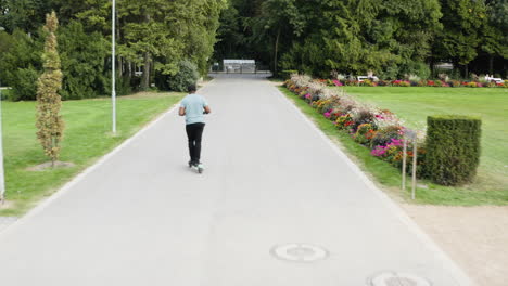 Rear-view-of-man-on-e-scooter-going-forward-on-pathway-park,-dolly-in,-day