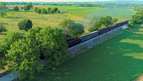 Luftaufnahme-Einer-Antiken-Dampflokomotive-Und-Personenwagen,-Die-Durch-Die-Landschaft-Fahren-Und-Rauch-Ausblasen,-Und-Einer-Drohne,-Die-An-Einem-Sonnigen-Sommertag-Vor-Ihr-Vorbeifliegt,-Während-Sie-Eine-Kleine-Brücke-überquert