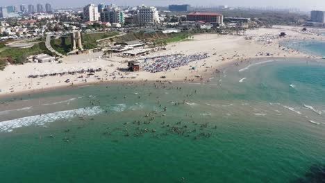 Several-tourists-and-locals-enjoy-the-warm-waters-of-the-Mediterranean-on-a-sunny-day-with-the-skyline-of-Herzeliya-in-Israel-in-the-background