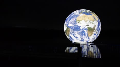 Luke-Jarram-Exposición-De-Arte-De-Tierra-Flotante-Iluminada-Reflejos-En-El-Agua-Del-Lago-Por-La-Noche-Descendiendo-A-La-Silueta-De-Barreras