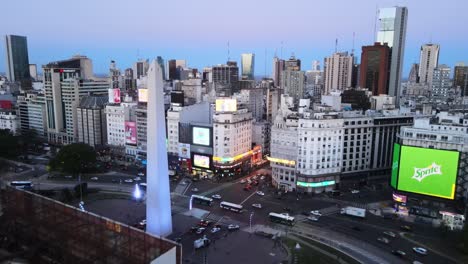 Aerial-pull-out-shot-from-Obelisco-monument-of-Buenos-Aires-capturing-traffic-motions-and-downtown-cityscape-with-flashy-billboards-display,-reveals-heritage-rooftop-chalet-muebleria-diaz-at-twilight