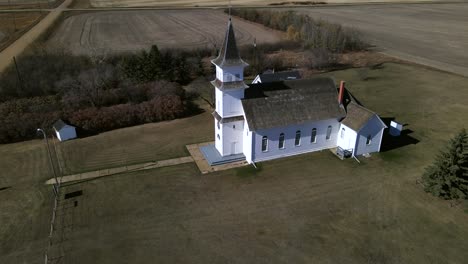 Drone-orbiting-around-old-country-church-in-Alberta's-prairies-while-slowly-ascending-and-revealing-young-person-walking-along-a-path-towards-the-building