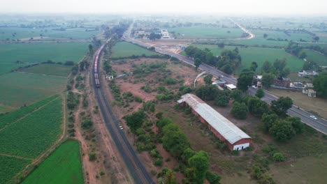 Freight-train-approaching-the-camera-with-aerial-drone-view,-the-wagons-are-coloured-blue,-yellow-and-red,-and-they-are-moving-on-the-tracks-next-to-a-highway-and-green-fields-in-India