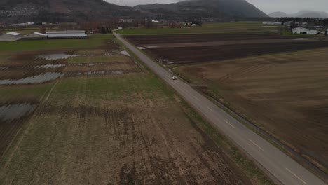 white-car-driving-along-country-farm-road-surrounded-by-grass-brown-fields-mountains-in-farmland-Abbotsford-BC-Aerial-tracking-orbit-wide-above