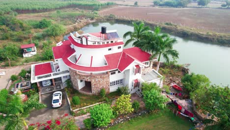Rotating-aerial-view-of-a-beautiful-complex-of-three-story-luxury-white-houses,-surrounded-by-a-private-forest,-palm-trees-and-a-boating-lake-for-relaxing-and-vacationing-in-Vadodara,-India