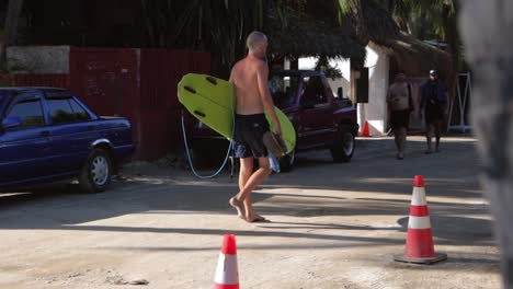 Kaukasischer-Männlicher-Surfer-Ohne-Hemd-Und-Grünem-Surfbrett,-Der-Barfuß-Auf-Einer-Schlammigen-Unbefestigten-Straße-In-Mexiko-Läuft,-Nachdem-Er-Wellen-Gefangen-Hat