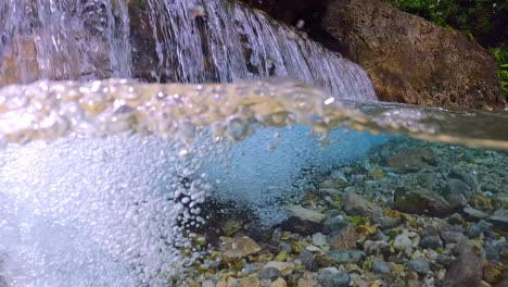 Close-up-slow-motion-underwater-shot-of-small-waterfall-floating-into-clear-river-with-rocks-on-the-ground