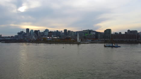 Boat-passing-on-river,-aerial-drone-view-of-old-port,-Montreal,-Quebec,-Canada,-downtown-in-background,-ferris-wheel,-tower