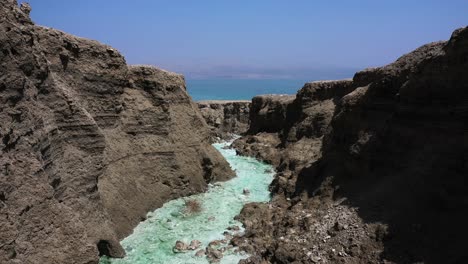Dead-Sea-Hot-Springs-dramatic-aerial-fly-in-through-desert-canyon-landscape-over-fast-running-crystal-clear-warm-waters-on-a-sunny-day