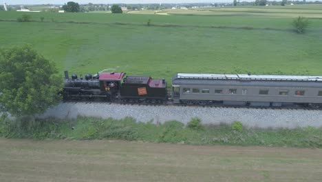 Vista-Aérea-De-Un-Motor-De-Vapor-De-1910-Con-Un-Tren-De-Pasajeros-Que-Echa-Humo-Viajando-A-Lo-Largo-De-La-Campiña-Amish-En-Un-Día-Soleado-De-Verano-Visto-Por-Un-Dron