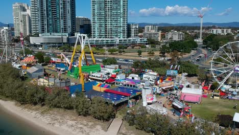 Aerial-view-of-a-beach-side-carnival-next-to-a-busy-main-road-with-buildings-in-the-background