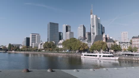 Static-shot-of-Frankfurt-Skyline-with-Skyscrapers-and-ferry-and-eiserner-Steg-Bridge-in-forefront,-view-from-main-river-on-a-sunny-day-with-clear-sky,-hessen,-Germany