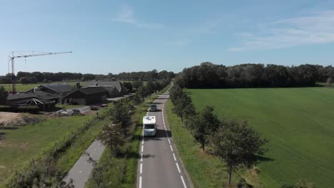 Seguimiento-Aéreo-De-Una-Caravana-En-Una-Carretera-Asfaltada-Limpia-En-Los-Países-Bajos-Con-Una-Hilera-De-árboles-Contra-Un-Cielo-Azul