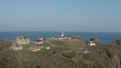 Aerial-pivot-shot-of-Gamov-lighthouse-building-complex,-standing-on-steep-rocky-cliff,-with-green-grass-hill,-on-the-sunset