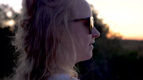 Young-blonde-woman-with-sunglasses-that-is-looking-to-beautiful-nature-at-sunset,-close-up