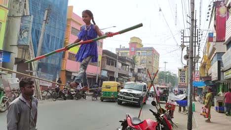 Poor-little-orphan-Indian-girl-balancing-long-stick-and-walking-on-rope-as-street-performer-at-daytime,-vehicles-in-background,-Slow-motion