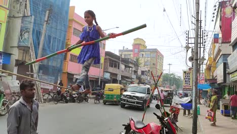 Pobre-Niña-India-Huérfana-Balanceando-Un-Palo-Largo-Y-Caminando-Sobre-Una-Cuerda-Como-Artista-Callejero-Durante-El-Día,-Vehículos-En-El-Fondo,-Cámara-Lenta