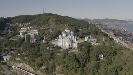 Toma-Aérea-De-Pivote-Lento-De-Una-Iglesia-Ortodoxa-Con-Techo-Azul-Y-Cúpulas-Doradas,-Ubicada-En-La-Cima-De-La-Colina-Con-El-Puerto-Y-El-Edificio-De-La-Ciudad-En-El-Fondo,-En-Un-Día-Brillante,-Claro-Y-Soleado