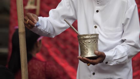 Un-Sacerdote-Hindú-Vestido-De-Blanco-Usa-Una-Pequeña-Flor-Para-Recoger-Agua-De-Una-Taza-Y-Rociarla-Sobre-Los-Creyentes-En-Una-Ceremonia-Al-Aire-Libre.