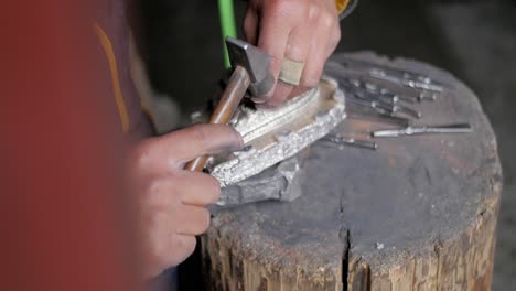 Eine-Erfahrene-Handwerkerin-Schnitzt-Sorgfältig-Mit-Hammer-Und-Meißel-Ein-Design-Auf-Einen-Kleinen-Silbernen-Hügel-Auf-Einem-Kleinen-Baumstamm-–-Nahaufnahme