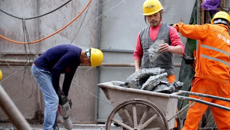 Construction-workers-breaking-concrete-surface-on-a-building-site-using-demolition-hammer-power-tool-and-a-wheelbarrow,-Slow-motion-handheld-shot