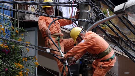 Two-utility-electrician-workers-fix-a-broken-voltage-cable-line-high-on-a-pole-on-the-street,-handheld-shot