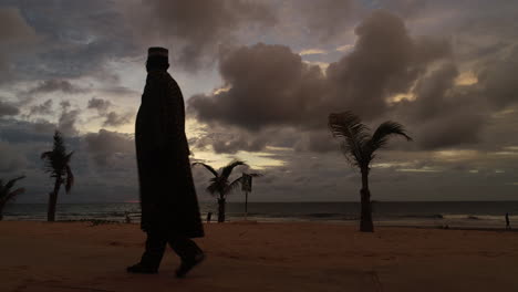 Ein-Afrikanischer-Mann-Mit-Dashiki-Geht-Bei-Sonnenuntergang-Von-Rechts-Nach-Links-Am-Strand-Von-Senegambia,-Serekunda,-Gambia-Vorbei