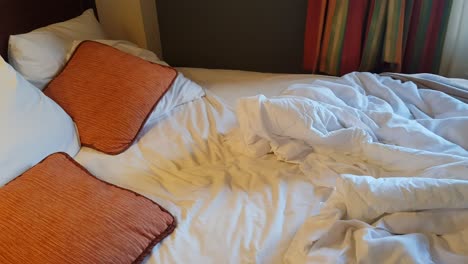 A-messy-hotel-bed,-with-white-sheets-and-orange-cushions
